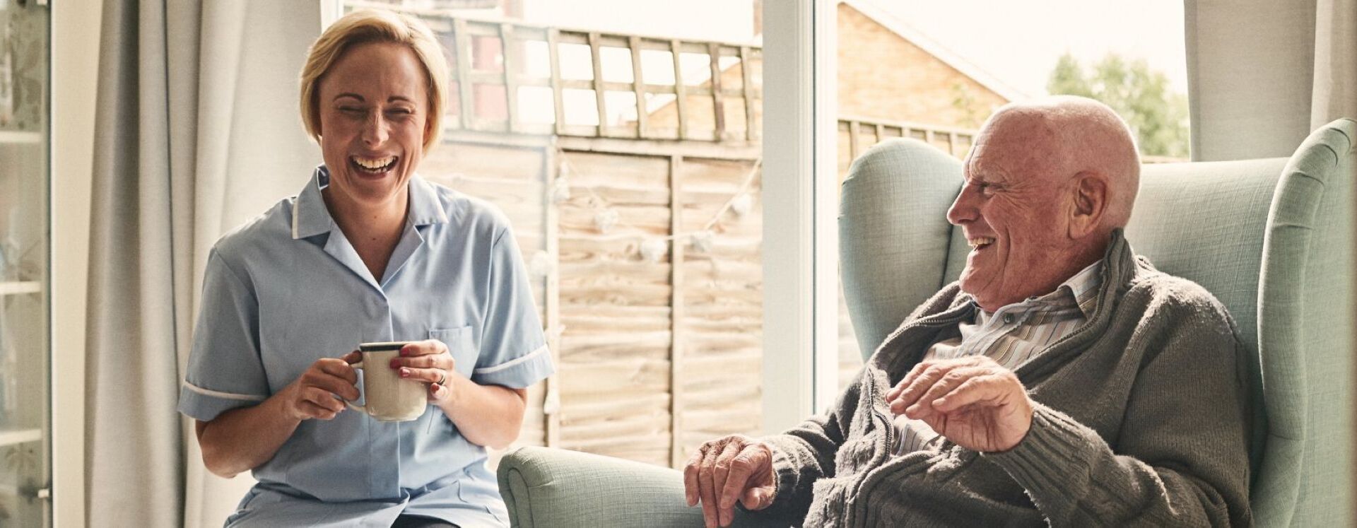 Man sitting and chair and woman sitting next to him laughing holding coffee